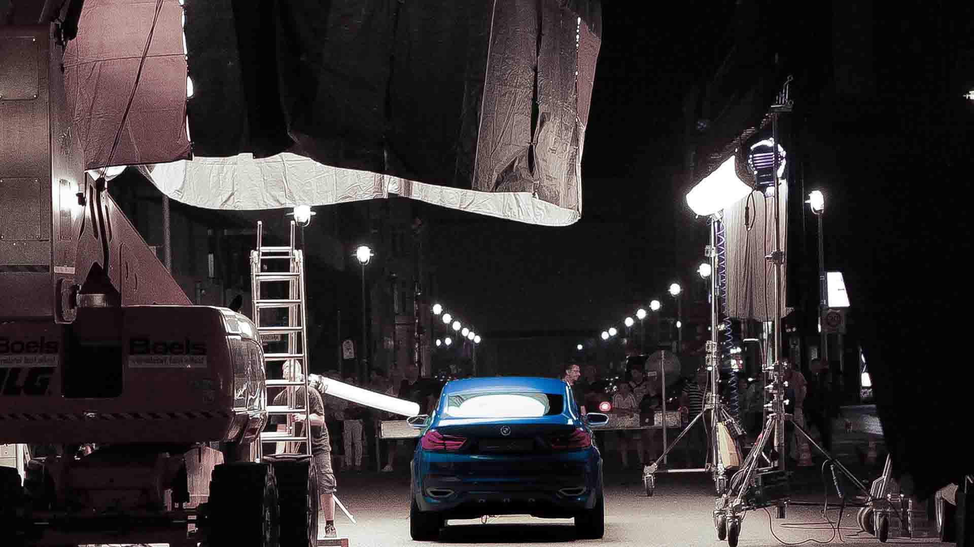 Das Bild zeigt ein parkendes Auto von hinten. Es steht in einer Halle. Es ist ein blauer BMW Concept X4. Um ihn herum stehen Lampen, Leuchten und eine Leiter. Das Bild dient als Making Of Bild für den Portfolioeintrag BMW Concept X4 von Panda Pictures.