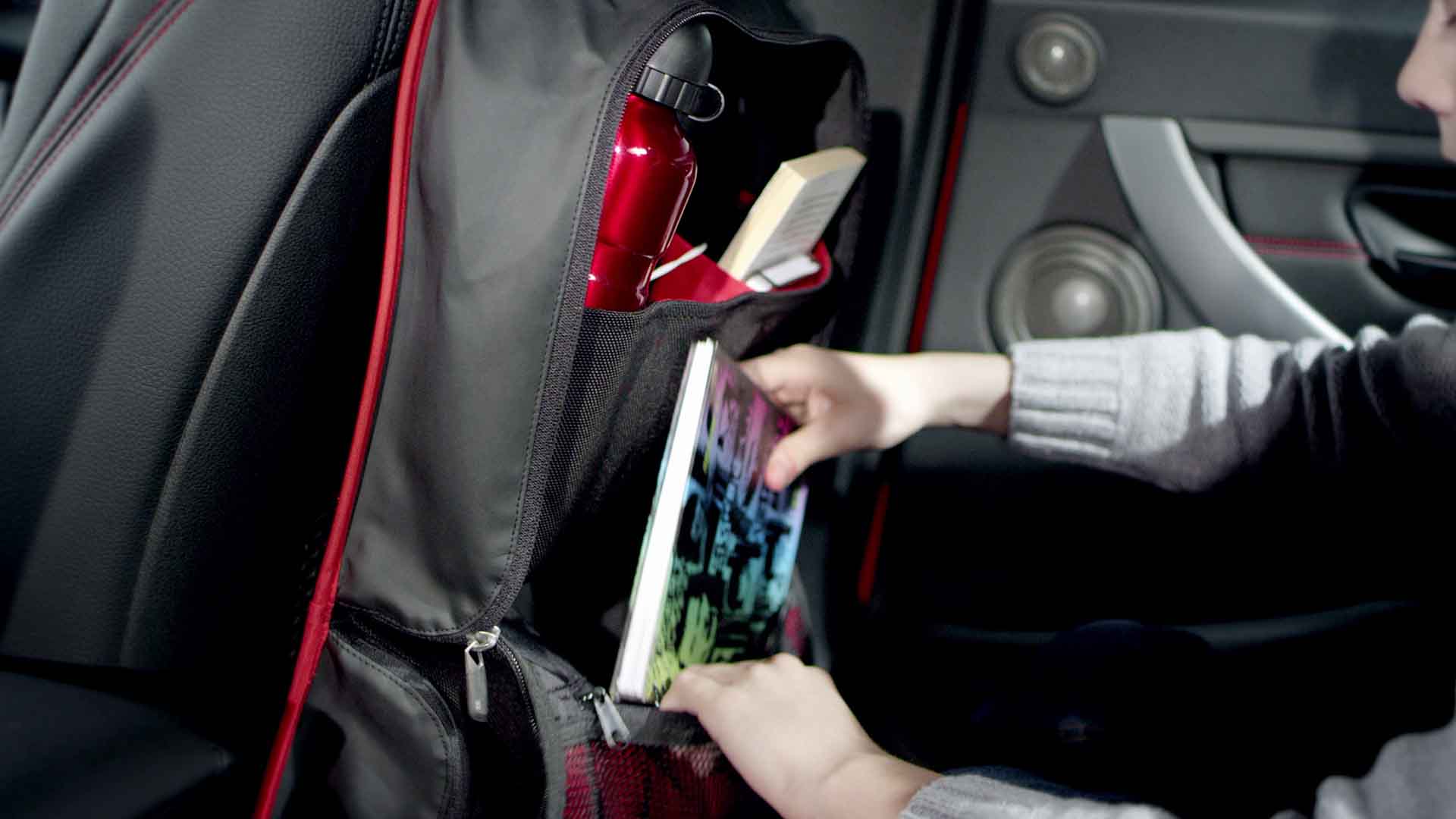 Das Bild zeigt eine Autotasche. Sie befindet sich am Rücksitz. Eine Kinderhand greift in die Tasche und holt ein Buch heraus. Die Tasche ist grau und besitzt rote Nähte. Das Bild dient als Sliderbild für den Portfolioeintrag BMW F30 Zubehör von Panda Pictures.