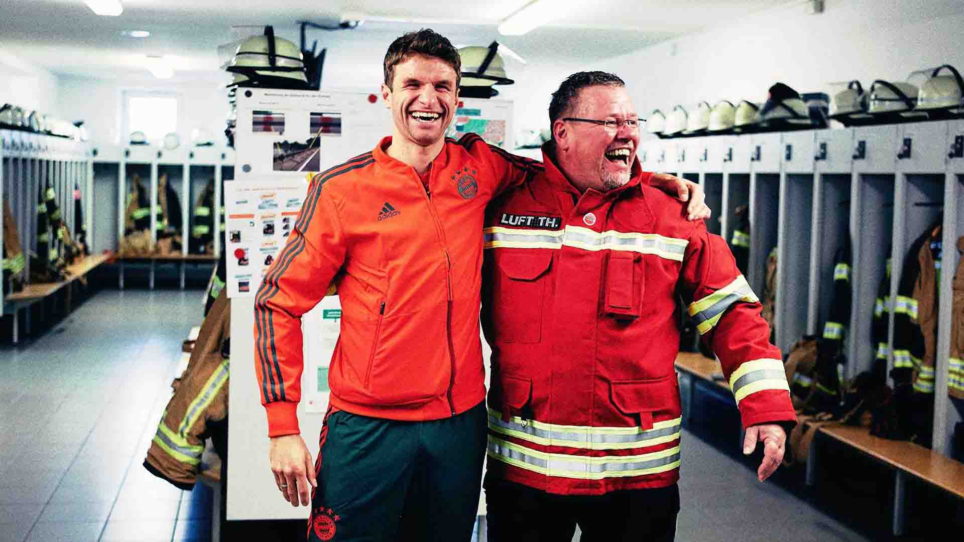 Das Bild zeigt Fußballstar Thomas Müller des FC Bayern Münchens und einen Feuerwehrmann. Beide Männer halten sich an den Schultern und lachen. Sie stehen in einer Umkleidekabine. Das Bild dient als Sliderbild für den Portfolioeintrag "Audi | FC Bayern München Ehrenrunde" von Panda Pictures.